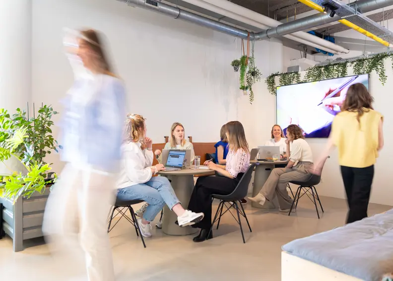 Moderne samenwerkingsruimte met jonge recruitment professionals in overleg aan laptops – creatieve werkomgeving met planten, designinterieur en digitale presentatie op scherm.