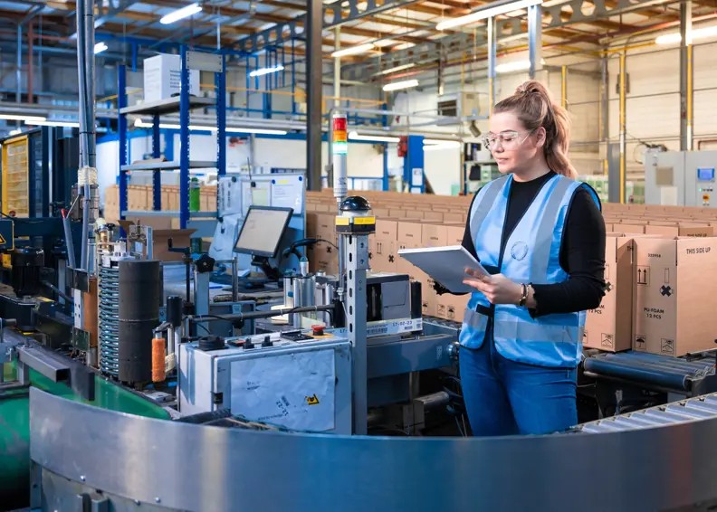 Vrouw Met Nrg Office Veiligheidsvest Aan In Een Magazijn Omgeving Met Kladblok In De Hand (1)