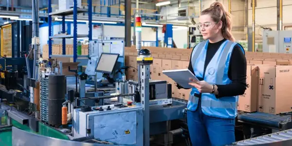 Vrouw Met Nrg Office Veiligheidsvest Aan In Een Magazijn Omgeving Met Kladblok In De Hand (1)